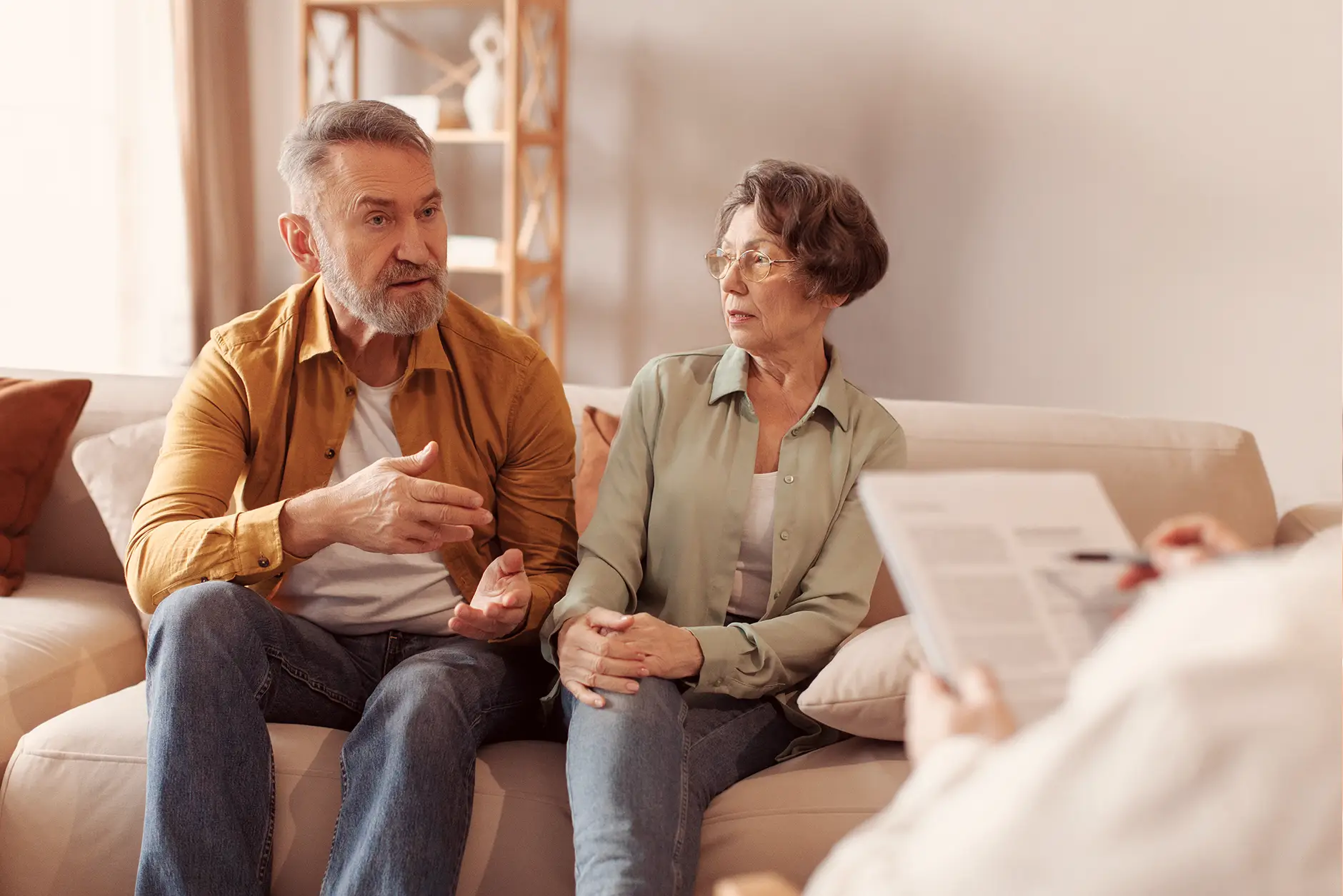 Ein älteres Paar sitzt auf einem Sofa und spricht aufmerksam mit einer dritten Person, vermutlich in einem Beratungsgespräch